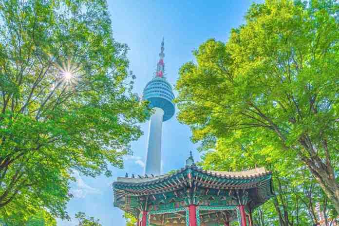 A view of a tall tower surrounded by lush green trees, with a traditional gazebo in the foreground under a clear blue sky.