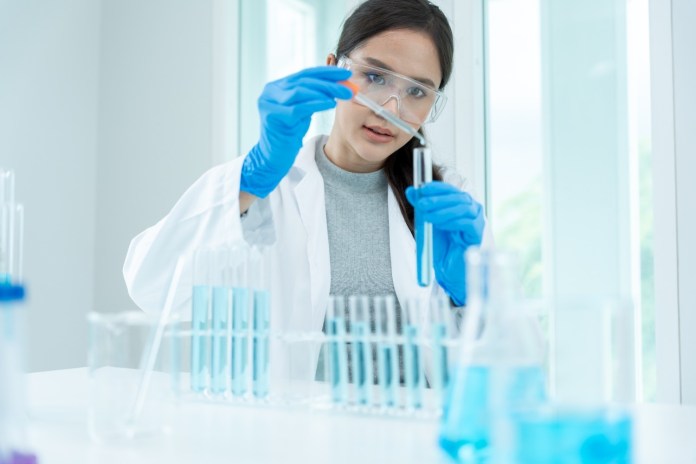 A woman in a lab coat and safety goggles is carefully measuring a liquid using a pipette in a laboratory setting, surrounded by various test tubes and glassware filled with blue liquid.