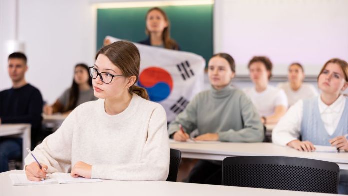 A classroom scene with a focus on a female student taking notes, wearing glasses and a white sweater, while other students appear engaged in the background. One student is holding a flag with a circular design.