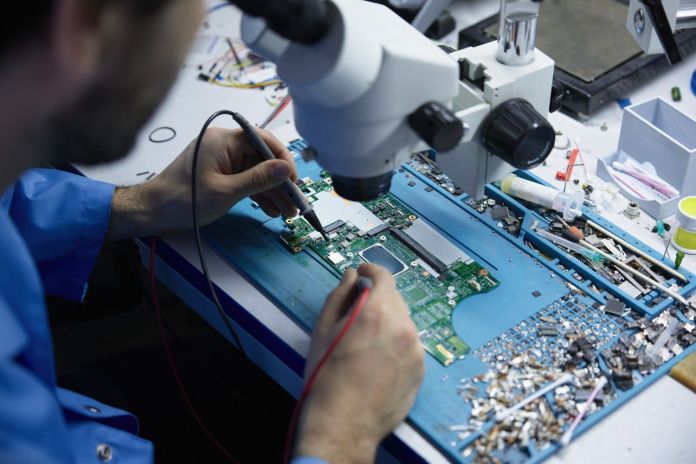 A technician working on a circuit board under a microscope, using a soldering tool in a cluttered electronic repair workspace.