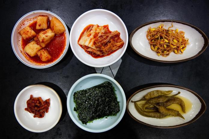 An assortment of Korean side dishes, including spicy radish, kimchi, bean sprouts, seaweed, and pickled green peppers, arranged in small bowls on a dark surface.