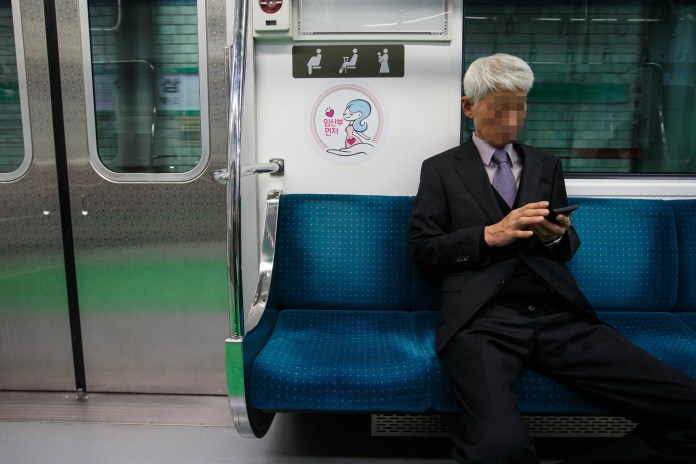 An elderly man in a suit sitting on a subway bench, looking at his phone, with blurred facial features for privacy. A sign depicting a cartoon character is visible on the wall beside him.
