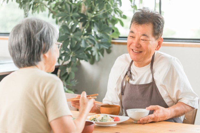 An older man and woman sitting at a table enjoying a meal together, smiling and engaged in conversation, with a plant in the background.