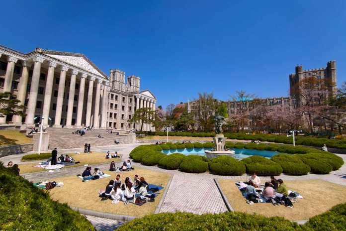 A vibrant campus scene featuring students relaxing on blankets in a green park area, surrounded by historic buildings and a fountain under a clear blue sky.