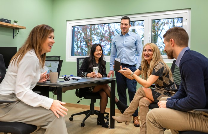 A group of four professionals engaged in a discussion in a modern office setting. Two women and two men are sitting around a table, with one woman showing something on her phone while others are actively listening and participating. The background features a green wall and a window with natural light coming in.