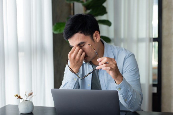 A man sitting at a desk with a laptop, rubbing his forehead in distress while holding a pair of reading glasses.