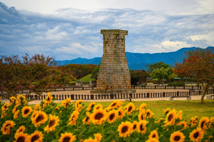 A historic stone tower surrounded by colorful sunflowers and lush greenery, with mountains and clouds in the background.