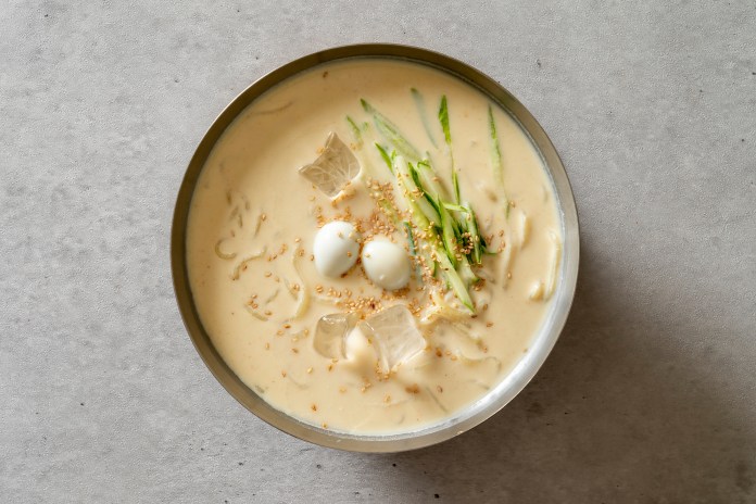 A bowl of creamy noodle soup topped with sliced cucumber, quail eggs, sesame seeds, and ice cubes on a light gray surface.