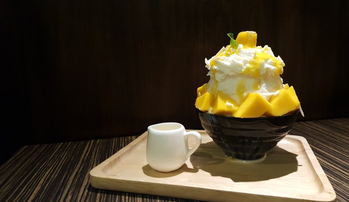 A bowl of mango shaved ice topped with whipped cream and mango chunks, placed on a wooden tray with a small jug of sweet sauce.