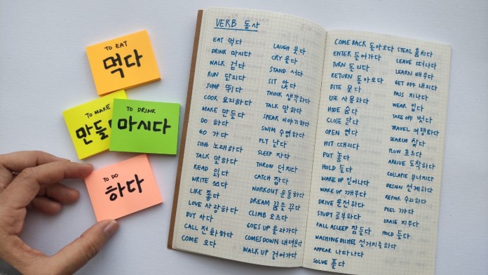 A hand holding sticky notes with Korean verbs: 'to eat' (먹다), 'to make' ( 만들다), 'to drink' (마시다), and 'to do' (하다), next to a notebook filled with additional Korean verbs.