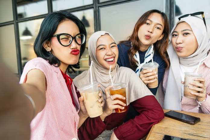 A group of four women posing for a selfie while holding drinks in a cafe, smiling and making playful expressions.