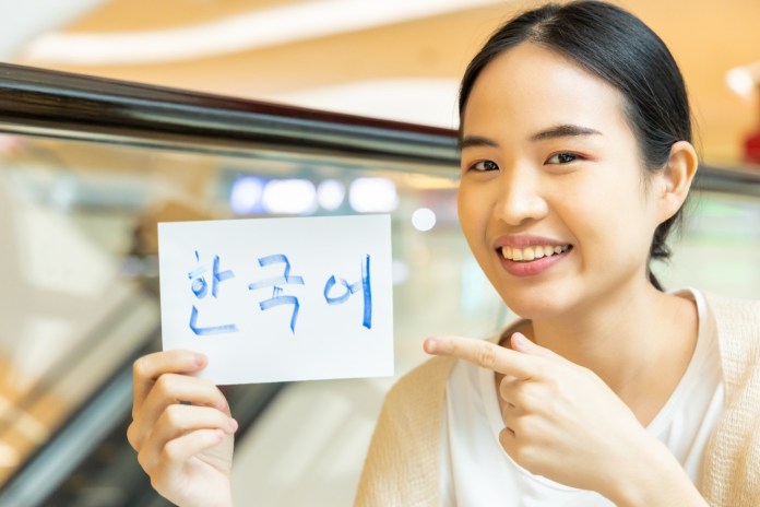 A woman smiling and pointing at a sign that reads '한국어' (Korean language), indoors in a shopping center.
