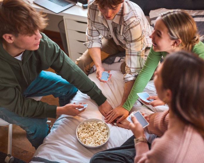 Four friends sitting on a bed playing a card game, with a bowl of popcorn in the center.