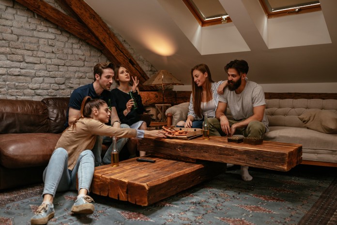 A group of five young adults sitting in a cozy living room with wooden beams and brick walls, playing a board game on a large wooden coffee table. Two people are focused on the game, while others are chatting and enjoying drinks.