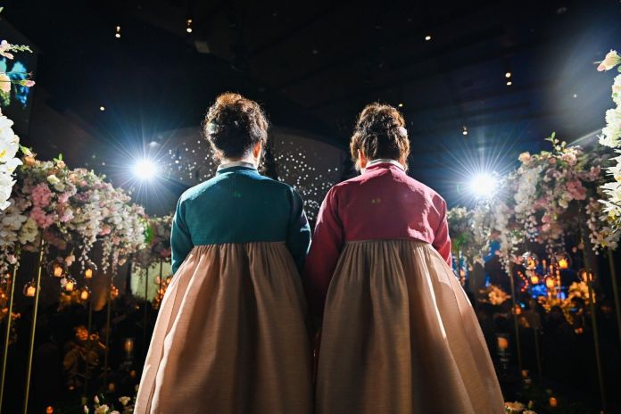 Two women in traditional Hanbok attire, seen from behind, standing among floral decorations in a dimly lit venue with bright lights.