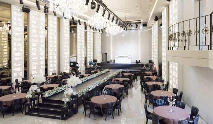Elegant event venue with round tables and black chairs, decorated with floral arrangements, featuring a stage and chandeliers.