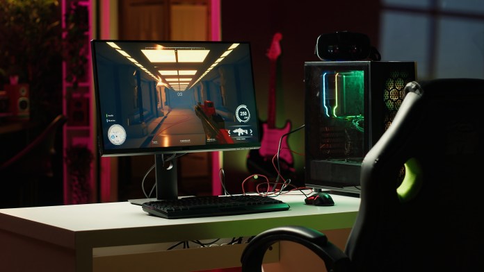 A gaming setup featuring a computer monitor displaying a video game, a keyboard, and a gaming mouse on a desk. A gaming chair is visible, along with a colorful PC case that has illuminated components.