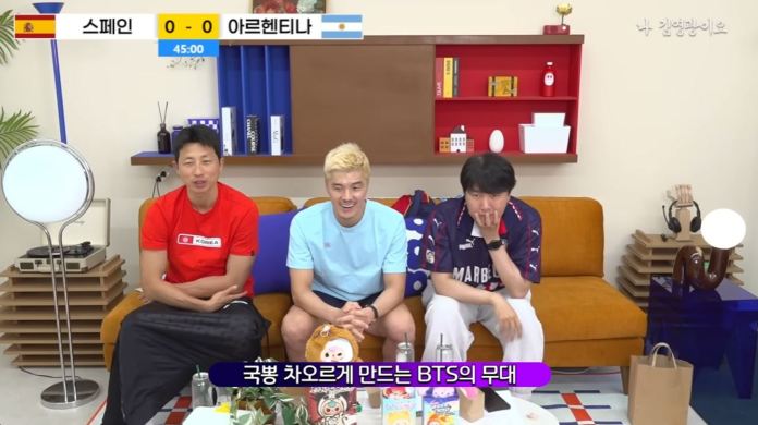 Three men sitting on a couch in a brightly colored room, watching a soccer game on a TV. The scoreboard shows Spain vs Argentina, 0-0 at halftime. One man is wearing a red shirt, another is in a blue shirt, and the third is in a navy jersey. Various decorative items are visible in the background.