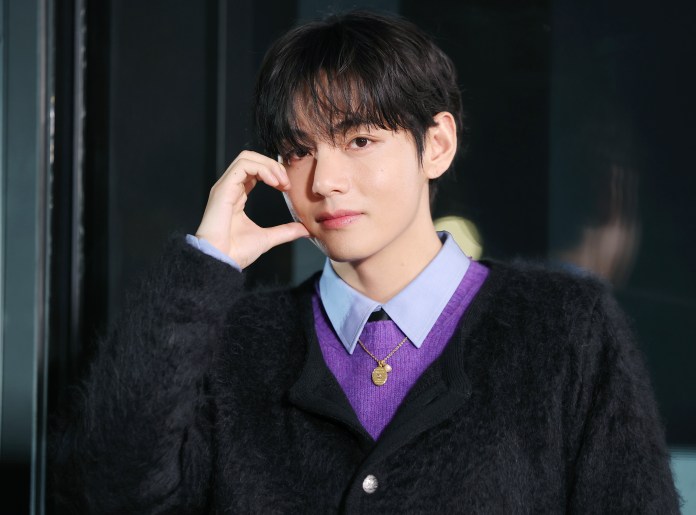 A young man with dark hair, wearing a black fuzzy sweater over a purple shirt, poses thoughtfully while resting his hand against his face.