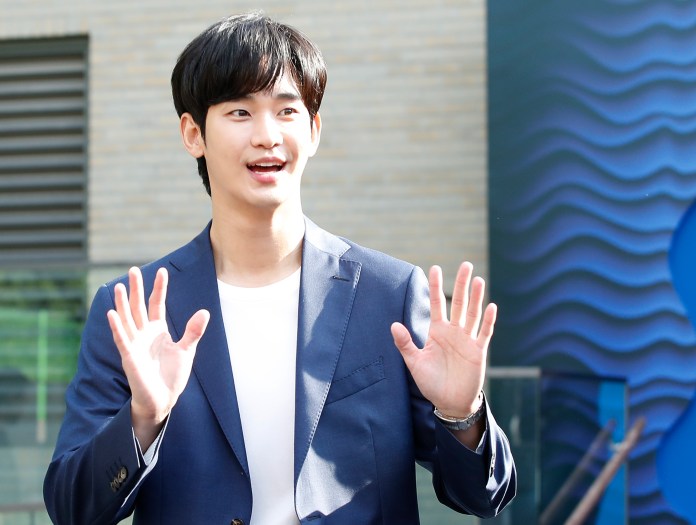 Kim Soo-hyun in a navy blue blazer and white shirt smiling and gesturing with both hands in a friendly manner.