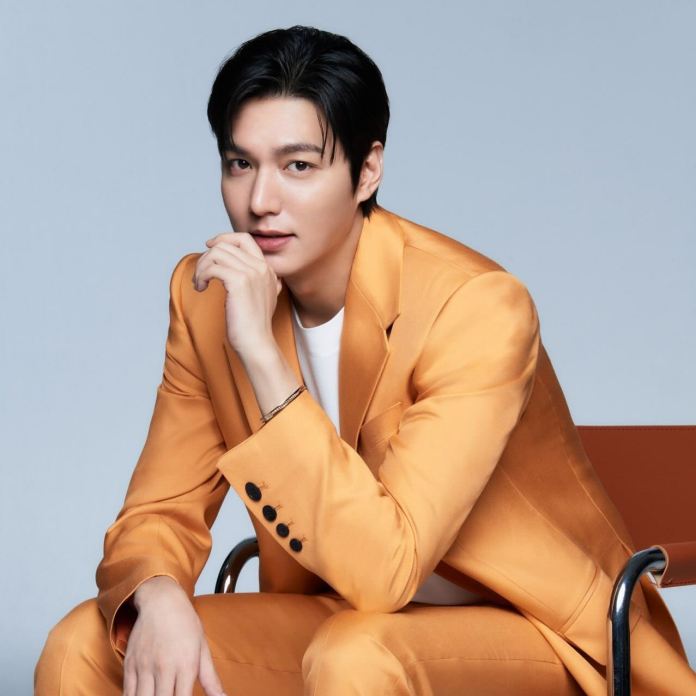 Lee min-ho in an orange suit, seated, with a contemplative expression, resting his chin on his hand, against a neutral background.