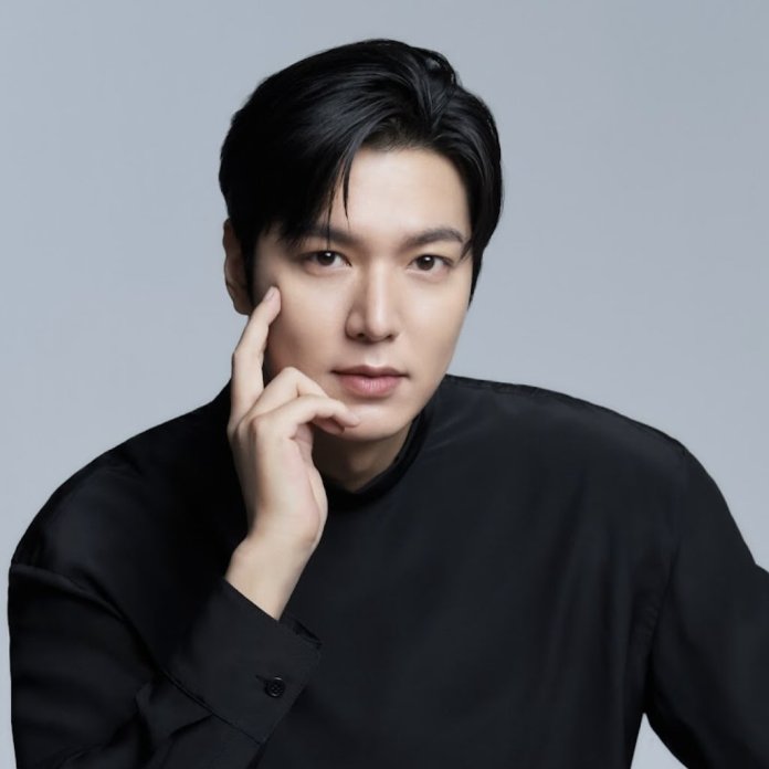 Close-up portrait of Lee min-ho with black hair, wearing a black shirt, resting his chin on his hand against a neutral gray background.