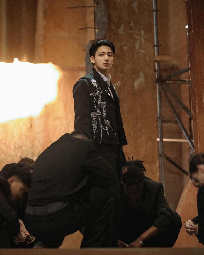 Jung Kook in a stylish black outfit with embellishments stands confidently among a group of people dressed in black, set against a rustic background with warm lighting.