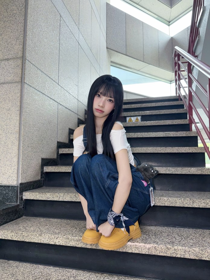 Woni sitting on stairs, wearing a white off-shoulder top and wide-leg denim pants, with a small plush toy visible on their side.