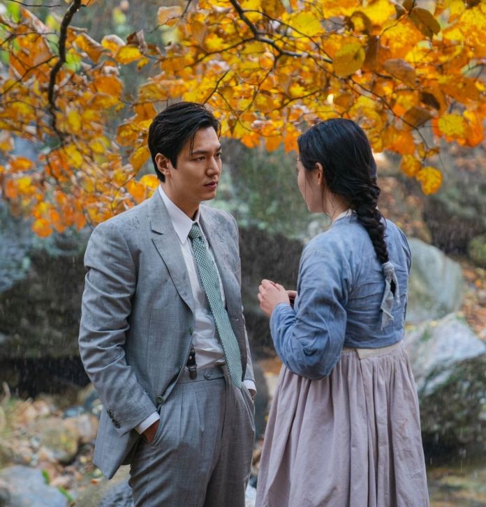 A man dressed in a gray suit and a woman in traditional attire share an intimate conversation under a backdrop of autumn leaves and a light rain.