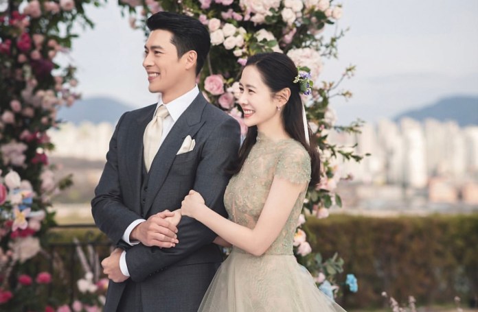 A smiling couple standing together in front of a floral backdrop, with the man dressed in a dark suit and the woman in a light, lacy gown, both looking happy and engaged.