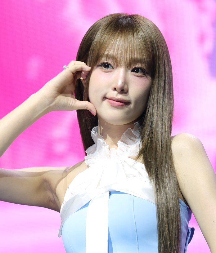 Choi Yena with long, straight hair posing with her hand near her face, wearing a light blue outfit with a ruffled neckline, against a pink background.