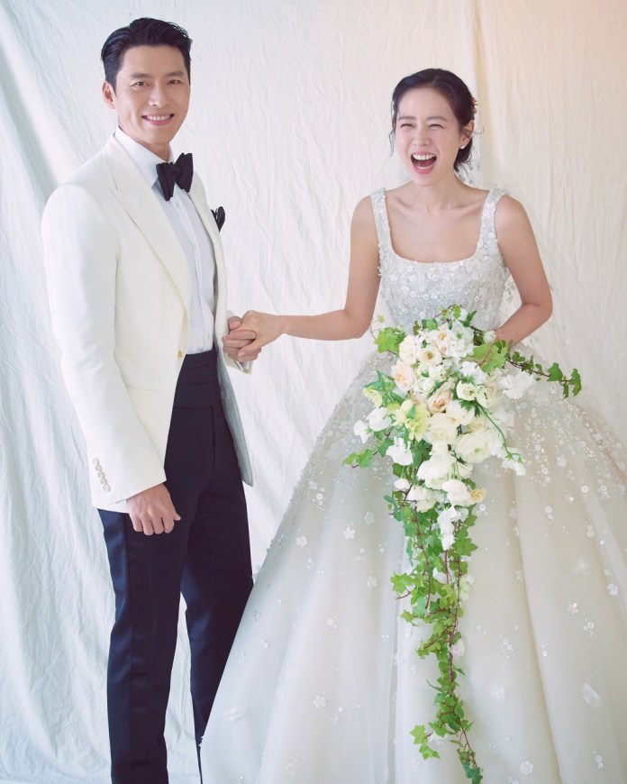 A smiling couple holding hands, dressed in formal wedding attire. The man is wearing a white jacket with a bow tie, and the woman is in a sparkling wedding gown adorned with flowers and greenery.