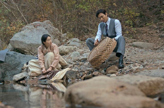 A man in a suit crouches by a stream, holding a woven basket, while a woman in traditional clothing sits nearby, looking surprised. The backdrop features rocky terrain and autumn foliage.