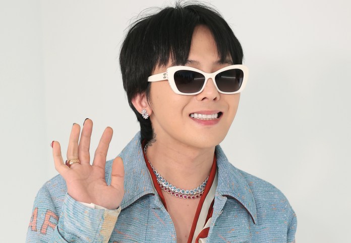A person wearing stylish sunglasses and a blue patterned jacket, smiling and waving, with accessories like a necklace and earrings.