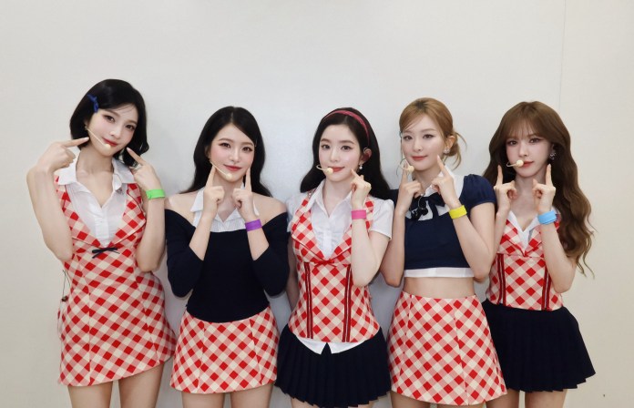 Red Velvet members wearing matching checkered outfits pose together, each making playful gestures with their fingers on their faces.