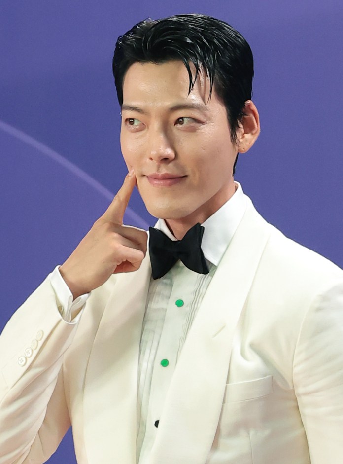 Kim Woo-bin in a white tuxedo and black bow tie poses playfully, touching his cheek and smiling against a purple background.