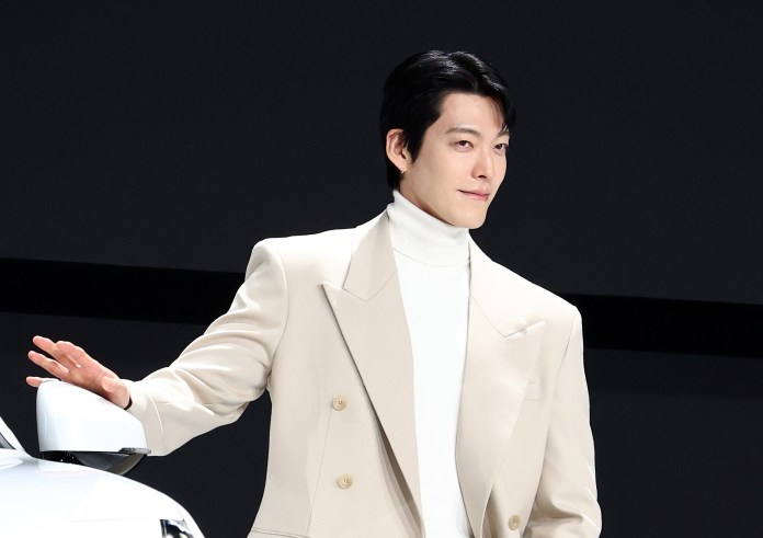 Kim Woo-bin in a white turtleneck and beige blazer poses next to a car, smiling confidently against a dark background.