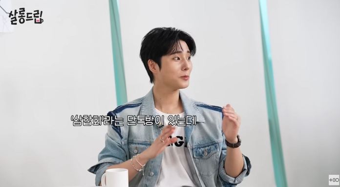 Young K with dark hair, wearing a denim jacket over a white shirt, gestures while speaking in a bright, modern setting.
