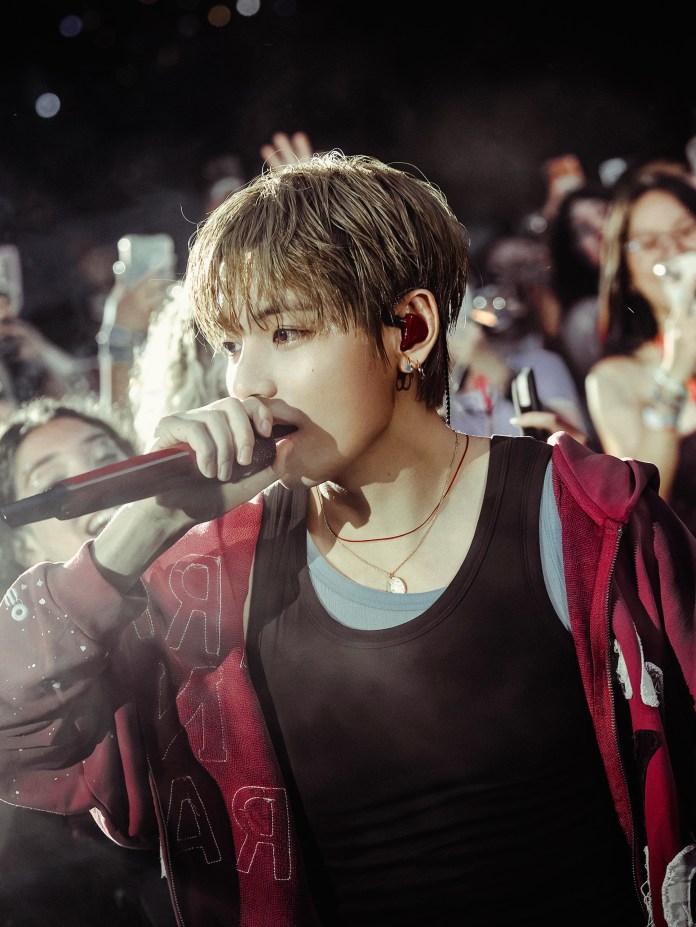 A close-up of a young male singer performing on stage, holding a microphone, with fans in the background and a focused expression.
