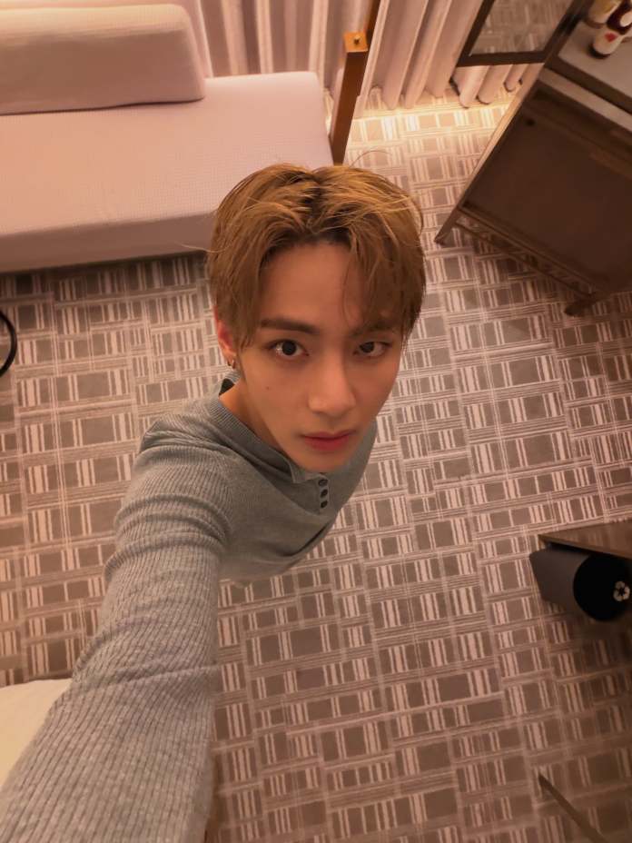 A person taking a selfie in a hotel room, wearing a gray long-sleeve shirt, with light brown hair and a neutral expression. The background features a patterned floor, a sofa, and a side table.