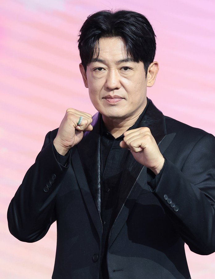 Man in a black suit posing with clenched fists and a serious expression, against a colorful background.