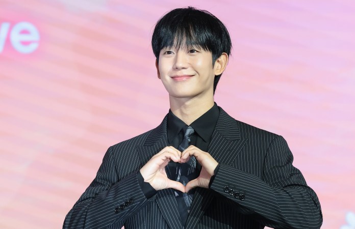 Jung Hae-in in a striped suit stands smiling at an event, making a heart shape with his hands against a colorful background.