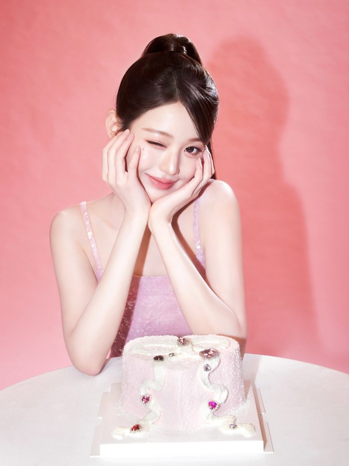 Jang Wonyoung in a light pink outfit winks while resting her cheeks on her hands, smiling at the camera, with a decorated pink cake on a table in front of her, against a pink background.