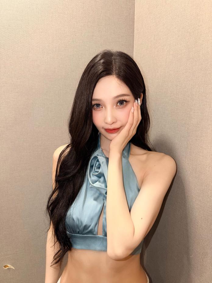 Joy with long dark hair, wearing a light blue halter top, holding her face with one hand against a neutral background.