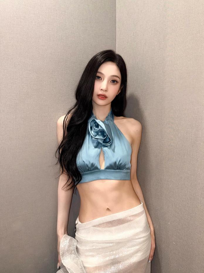 Joy posing against a gray wall, wearing a light blue halter top with a floral accent and a sheer white skirt.