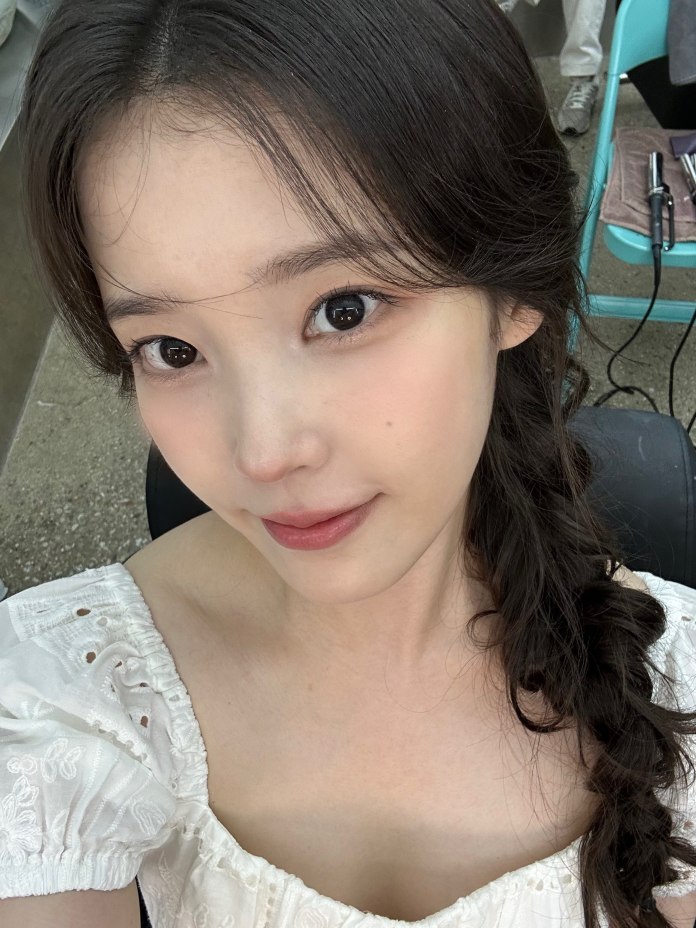 A close-up selfie of a young woman with long black hair styled in a loose braid. She is wearing a white off-the-shoulder top and has a natural makeup look, smiling softly at the camera.