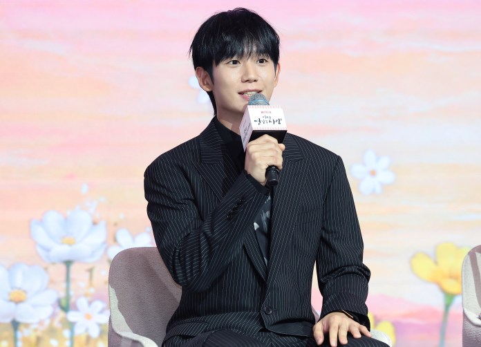 A man in a pinstripe suit sitting on a chair, holding a microphone and speaking at an event with a colorful floral background.