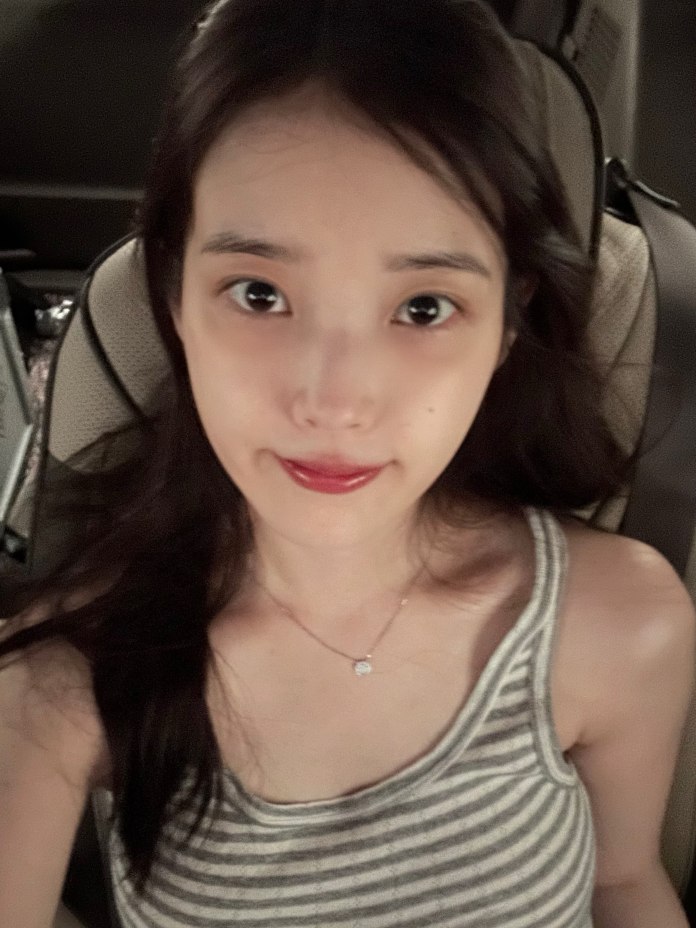 A young woman with long dark hair and a striped tank top is sitting in a car, looking directly at the camera with a slight smile.