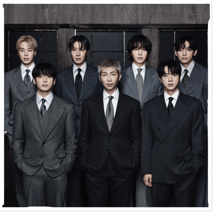 A group of eight men dressed in stylish suits, standing in two rows against a dark background.