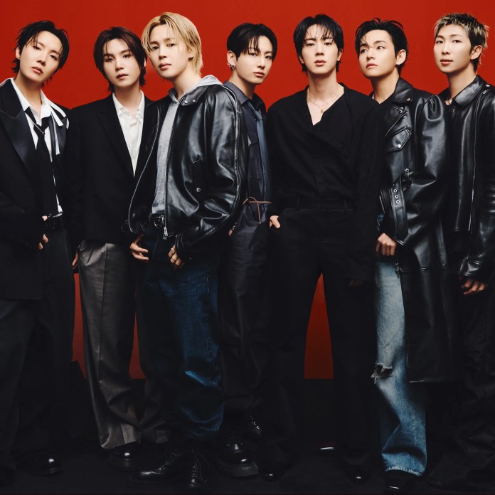 A group of seven individuals posing together against a red background, dressed in stylish, modern outfits, showcasing a mix of black leather, blazers, and denim. Their expressions are confident and serious.